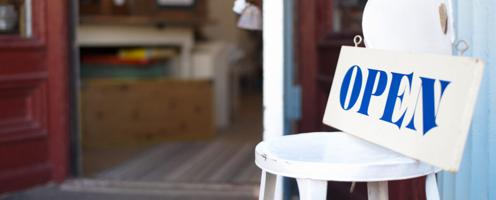 Hand-lettered wooden open sign sitting on chair in front of a downtown storefront with it's door open.